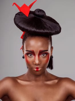 Portrait, makeup and black woman with origami for beauty in studio isolated on gray background. Face, creative cosmetics and confident model with red paper bird in hair, skincare and Japanese art.
