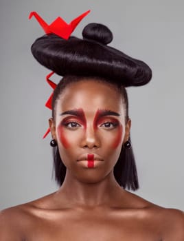 Portrait, cosmetics and black woman with origami for beauty in studio isolated on gray background. Face, creative makeup and confident serious model with red paper bird in hair, skincare and art.