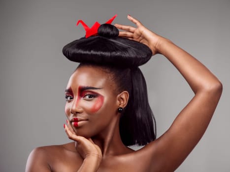 Portrait, makeup and black woman with origami in hair in studio isolated on gray background. Face, creative cosmetics and confident serious model with red paper crane for beauty, skincare and art.