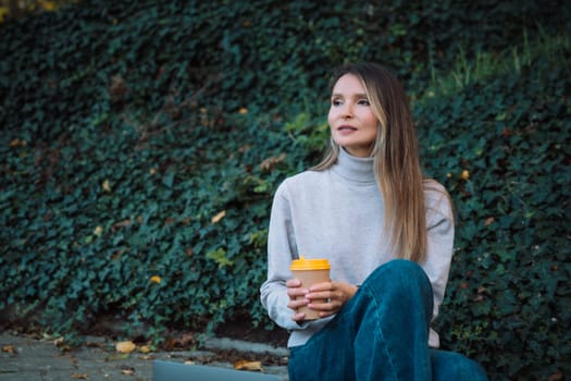 A woman sits on the ground with a cup in her hand. She is wearing a gray sweater and blue jeans