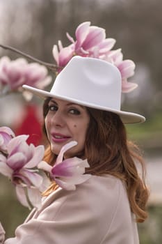 Woman magnolia flowers, surrounded by blossoming trees, hair down, white hat, wearing a light coat. Captured during spring, showcasing natural beauty and seasonal change