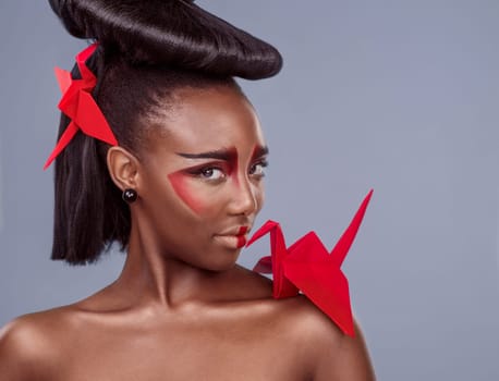 Portrait, cosmetics and black woman with origami for beauty in studio isolated on gray background. Face, creative makeup and confident serious model with red paper crane in hair, skincare and art.