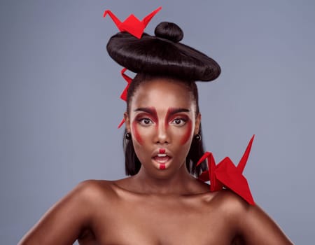 Portrait, makeup and surprise of black woman with origami for beauty in studio isolated on gray background. Shocked face, creative cosmetics and model with paper crane in hair, skincare and art color.