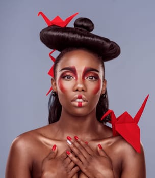 Portrait, makeup and black woman with origami for beauty in studio isolated on gray background. Face, creative cosmetics and confident young model with oriental paper crane in hair, skincare and art.