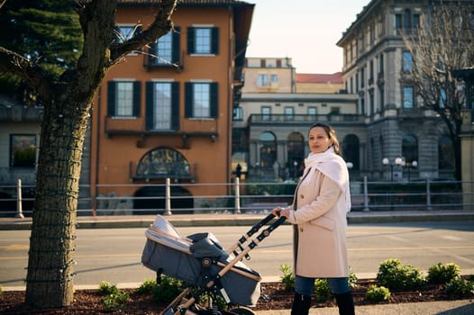Attractive Caucasian woman, loving caring mother in warm clothes enjoys a walk with her baby sleeping in baby carriage, admiring the beautiful city of Como in Italy. People, lifestyle and tourism