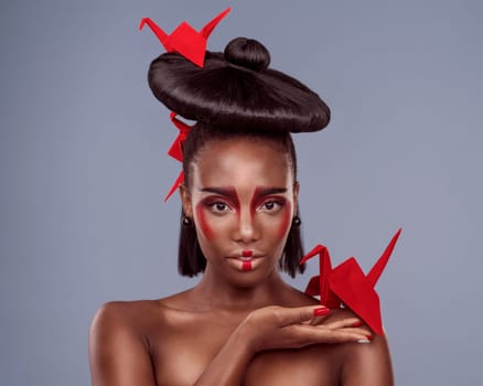 Portrait, cosmetics and black woman with origami for art, beauty or skin in studio isolated on gray background. Face, creative makeup and confident or serious model with oriental paper crane in hair.