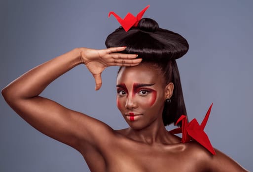 Portrait, beauty and black woman with origami for art, makeup or skin in studio isolated on gray background. Face, creative cosmetics and confident model with oriental paper crane in hair for culture.
