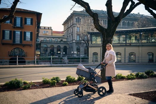 Young mother walking with her baby in stroller in the city on sunny winter day. Elegant pretty woman in warm beige winter coat, pushing baby pram while strolling long the city
