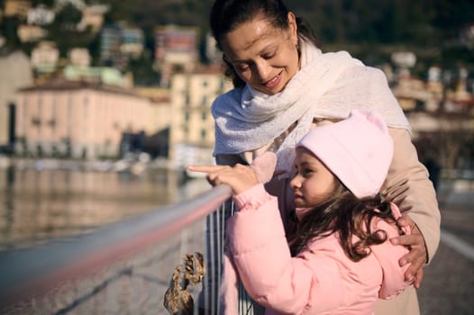 Happy loving mother and adorable daughter in warm clothes, enjoying beautiful view of the Como lake on windy sunny winter day. People. Leisure activity. Outdoor recreation. Travel and tourism concept