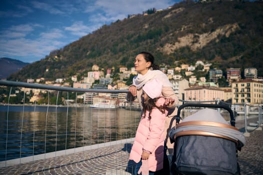 Happy young mother pushing stroller with little child, walking with her daughter outdoors, near a lake on sunny day. Happy family walking by the lake of Como in Italy. People. Travel. Lifestyle