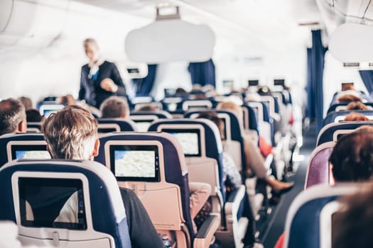 Interior of airplane with passengers on seats and stewardess in uniform walking the aisle, serving people. Commercial economy flight service concept