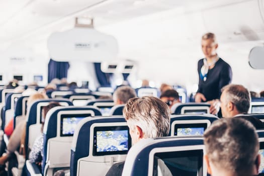 Interior of airplane with passengers on seats and stewardess in uniform walking the aisle, serving people. Commercial economy flight service concept