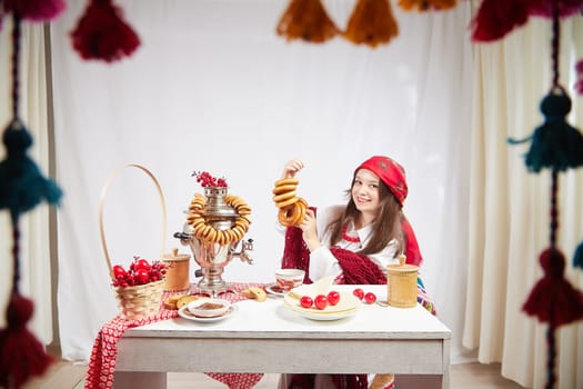 A fashionable modern girl in stylized folk clothes at table with a samovar, bagels and tea for the Orthodox holiday of Maslenitsa and Easter. Funny photo shoot for a young woman