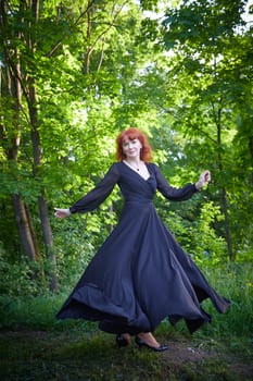 Elegant senior mature Woman dancing in Black Dress in green park. Woman with red hair posing on nature