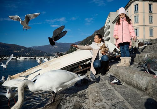 Young mother and her little daughter feeding swans at the Como lake, sitting against small boats over mountains background on a sunny winter day. Blue clear sky on background
