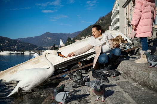 Young young woman, a mother and her little daughter feeding swans at the Como lake, sitting against small boats over mountains background on a sunny winter day. Blue clear sky on background