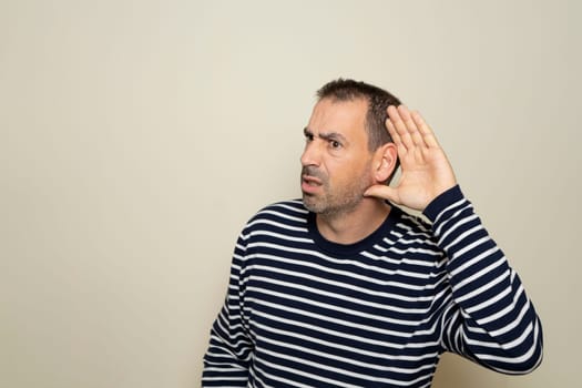 Hispanic man with beard in his 40s wearing a striped sweater listening to rumors keeping hand near ear isolated over beige color background