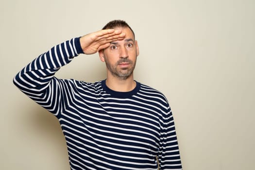 Hispanic man in his 40s saluting the camera with a military salute in an act of honor and patriotism, showing respect. Isolated on beige background