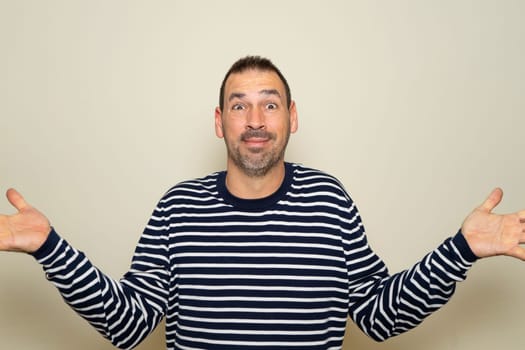 Hispanic man with beard in his 40s wearing a striped sweater clueless and confused with open arms, no idea concept. Isolated on beige studio background