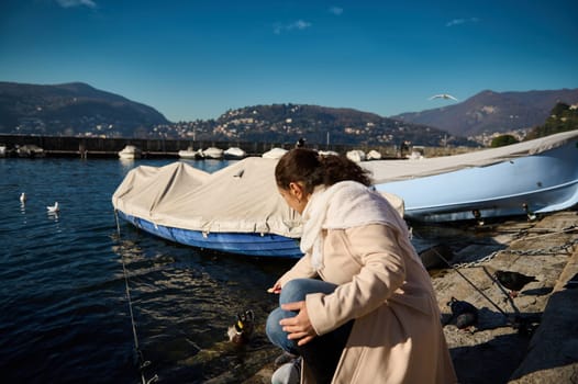 Rear view of a young woman in beige coat, sitting nears boats by the Como lake, relaxing in the lakefront and feeding by hands the birds and ducks with bread. People, nature and wild animals