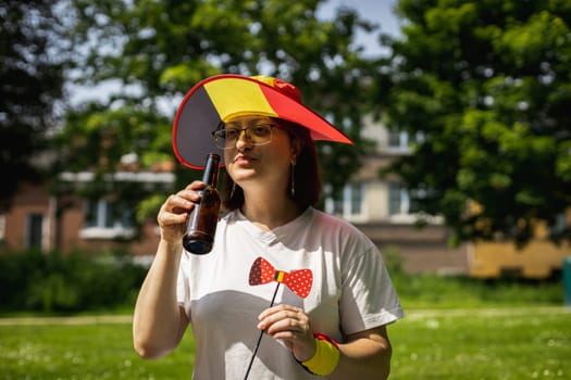 Portrait of a beautiful young caucasian woman in a hat drinking beer from a bottle and holding a paper bow decoration in her hand, celebrating belgium day at a picnic in a city park on a sunny summer day, close-up side view.