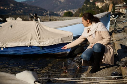 Young pretty woman crouching by ducks and swans on the lake of Como, feeding birds with bread, relaxing on the lakefront on a sunny and cold winter day. The province of Milan, Como, Lombardy, Italy