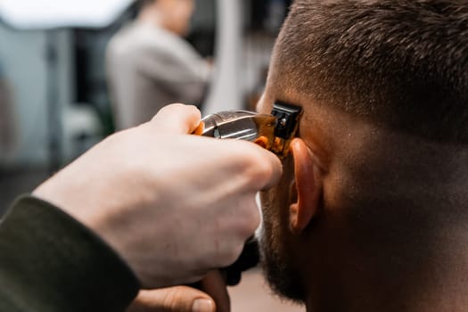 Skilled barber uses automatic trimmer to cut client hair on temple in barbershop closeup. Hairdresser does stylish hairdo by salon mirror