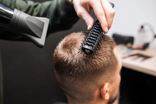 Barber brushes man short hair using dryer in barbershop. Skilled barber does elegant hairstyle in beauty salon. Male haircut