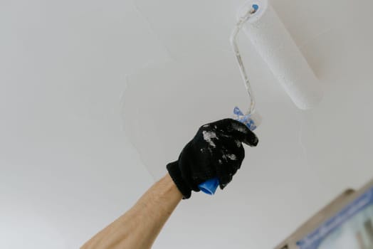 One black-gloved hand of a young unrecognizable Caucasian man paints the ceiling with white paint with a roller on a summer day in the kitchen, bottom side close-up view with selective focus.