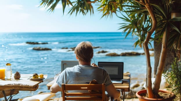 A man working on a laptop by the sea, surrounded by nature, under a clear sky