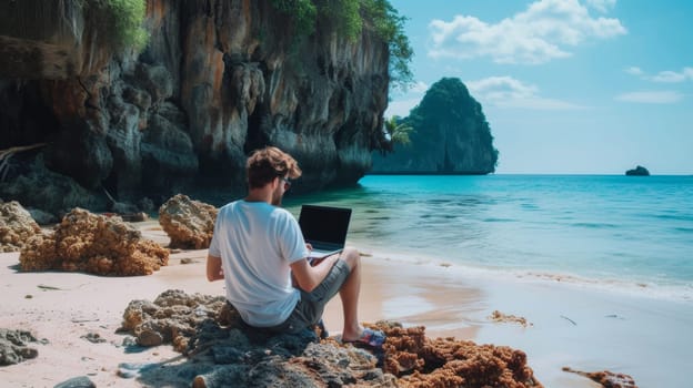 Individual working on a laptop seated on a sandy beach with a dramatic rocky landscape in the distance