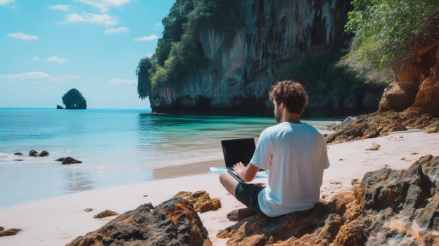 Individual working on a laptop seated on a sandy beach with a dramatic rocky landscape in the distance