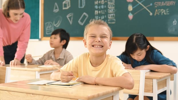 Caucasian smart child smiling at camera while doing classwork at classroom while happy multicultural student doing test or writing note in classroom at elementary class. Education concept. Pedagogy.