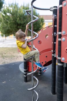 A young boy is climbing a spiral slide. on a children's playground The slide is red and black. The boy is wearing a yellow shirt