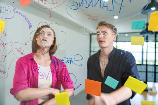 Couple of businesspeople discussing and brainstorming about solving marketing problem in front of glass board at business meeting by using mind map and sticky notes. Working together. Immaculate.