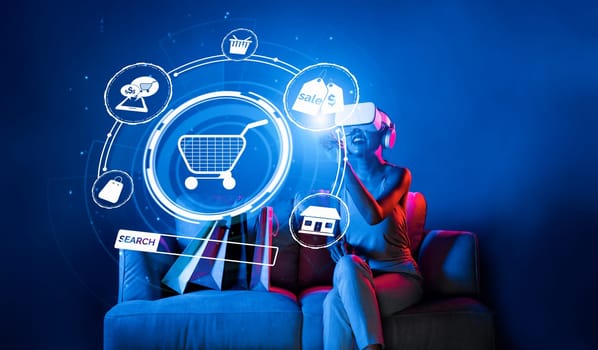 Smart female sitting on sofa while wearing VR headset connecting metaverse, future cyberspace community technology. Elegant woman enjoy open interface online store menu shop product. Hallucination.