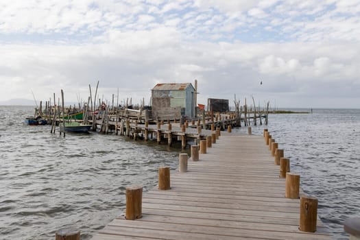 A pier with a wooden walkway leading to a small house. The pier is full of boats and the water is calm