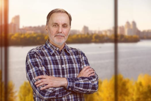 Portrait of a handsome confident mature man with folded arms. Blurred windows background with river scape view.