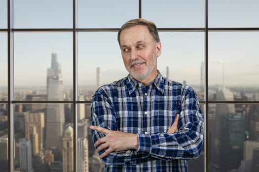 Cheerful mature senior man with folded arms standing in office pointing at copy space. Checkered windows background with cityscape view.