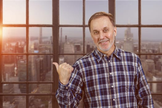 Portrait of smiling mature man pointing back with his thumb up. Checkered windows background with cityscape view.
