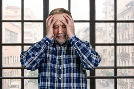 Portrait of stressed senior man holds his head. Elder mature man with headache standing indoors. Checkered windows background with town view.