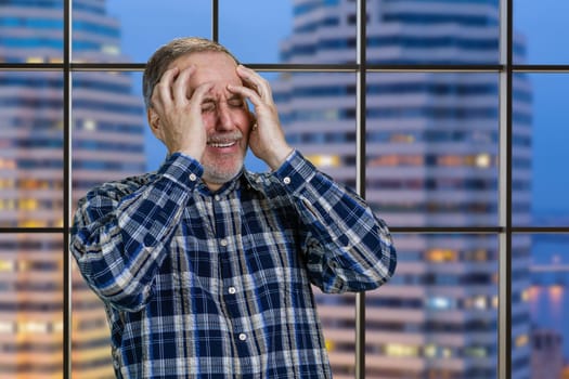Handsome mature man holds his head with both hands. Checkered windows background with night city view.