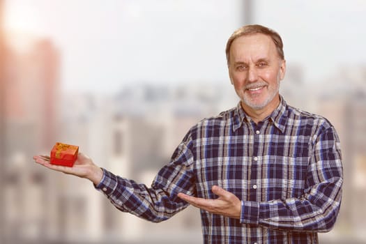 Happy senior man presenting a red gift box on his hand. Blurred cityscape background.