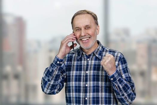 Happy cheerful senior man is talking on phone celebrating success. Blurred urban scape background.