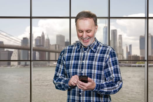 Portrait of a mature senior man is typing a message on his smartphone. Checkered windows background with view on a city river and skyscrappers.