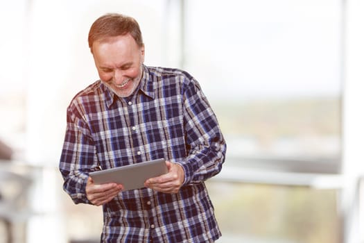 Happy mature aged man is looking down at tablet pc. Bright office background.