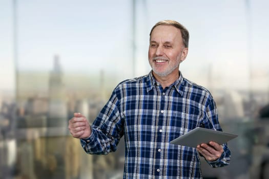 Happy handsome cheerful mature man with tablet pc. Indoor background with cityscape view.