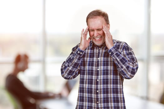 Portrait of mature man suffering from headache touching his temples with both hands. Bright office enviroment background.