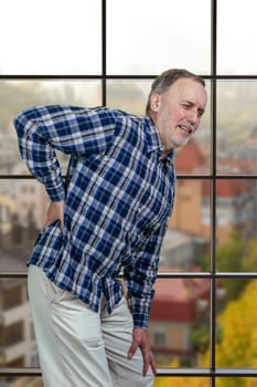 Old aged mature man is suffering from back pain. Checkered windows background. City scape view.