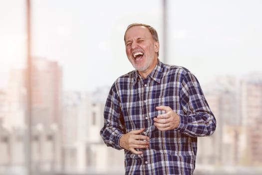Aged mature businessman is laughing hard out loud standing in office. Blurred office windows background.